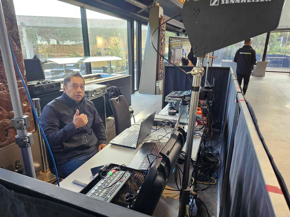 Manaia Go operator at a live streaming desk with vision switcher and laptops, beside carved pou at a Whānau Ora event