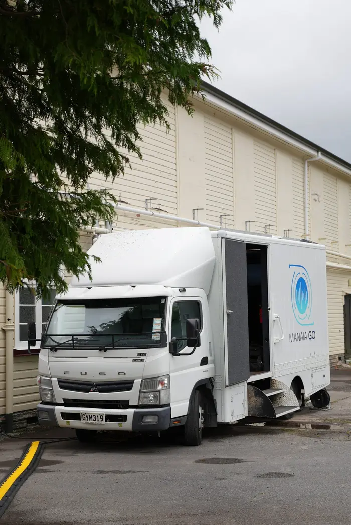 The white Manaia Go OB truck with its logo on the side, parked on site beside a hall with cables run out